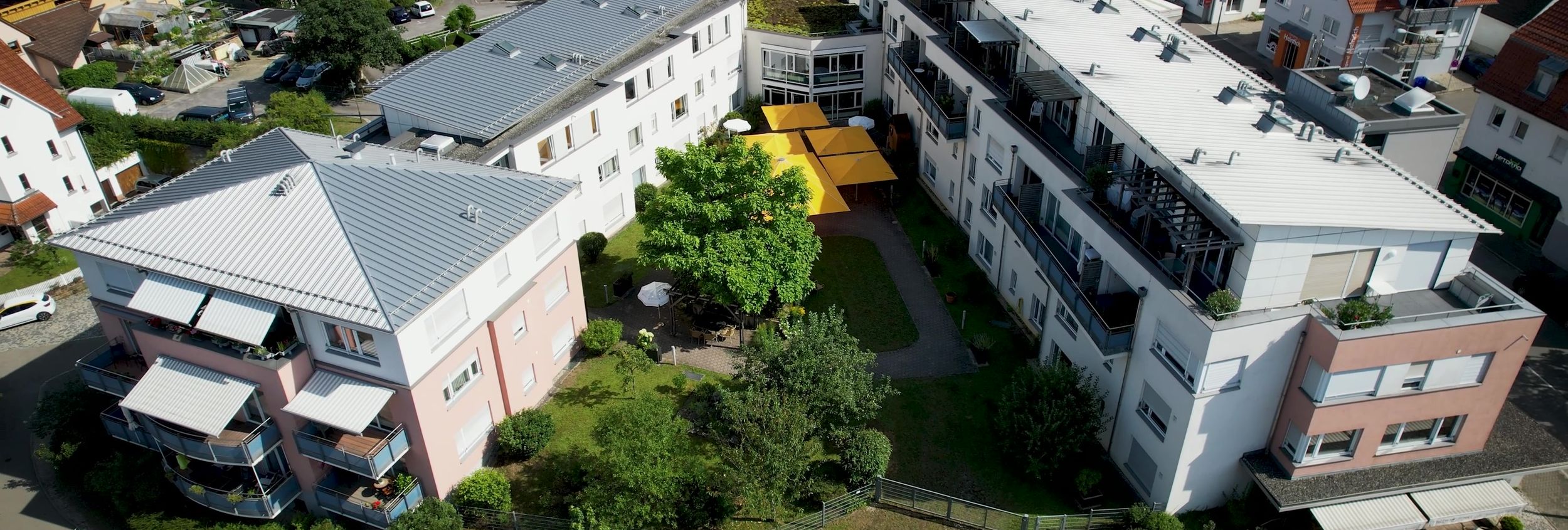 Luftaufnahme mehrerer Wohnhäuser um Innenhof mit gelben Sonnenschirmen